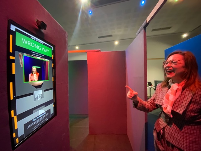 A woman laughs and points at a wall-mounted screen in a pink-walled maze corridor.