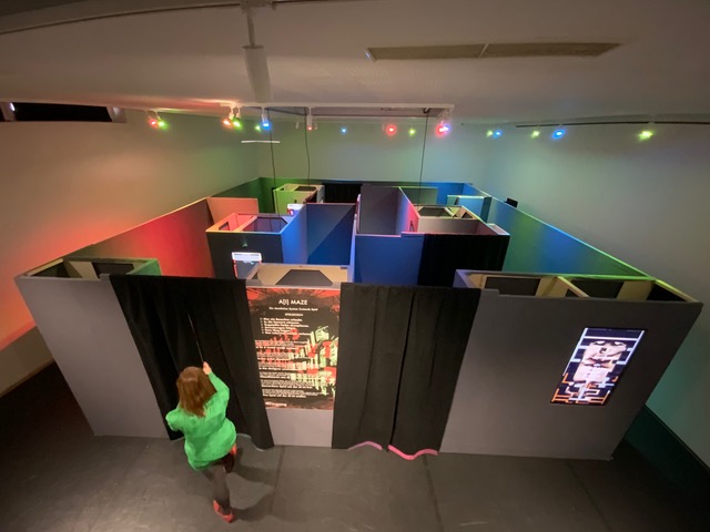 A person in a green hoodie stands beside a maze of black partitions lit with colorful lights.