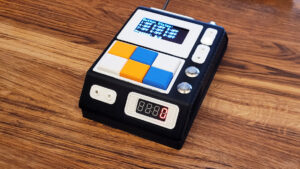 A small black tabletop device with a screen, colored square buttons, and side knobs sits on a wooden surface.