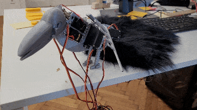 Partially assembled animatronic raven on a table, with exposed wires, servos, and black feathers beside a laptop.