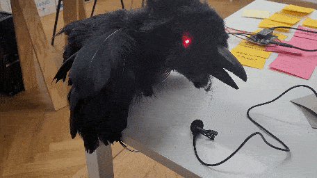 Black animatronic raven with glowing red eye and attached cable on a tabletop.