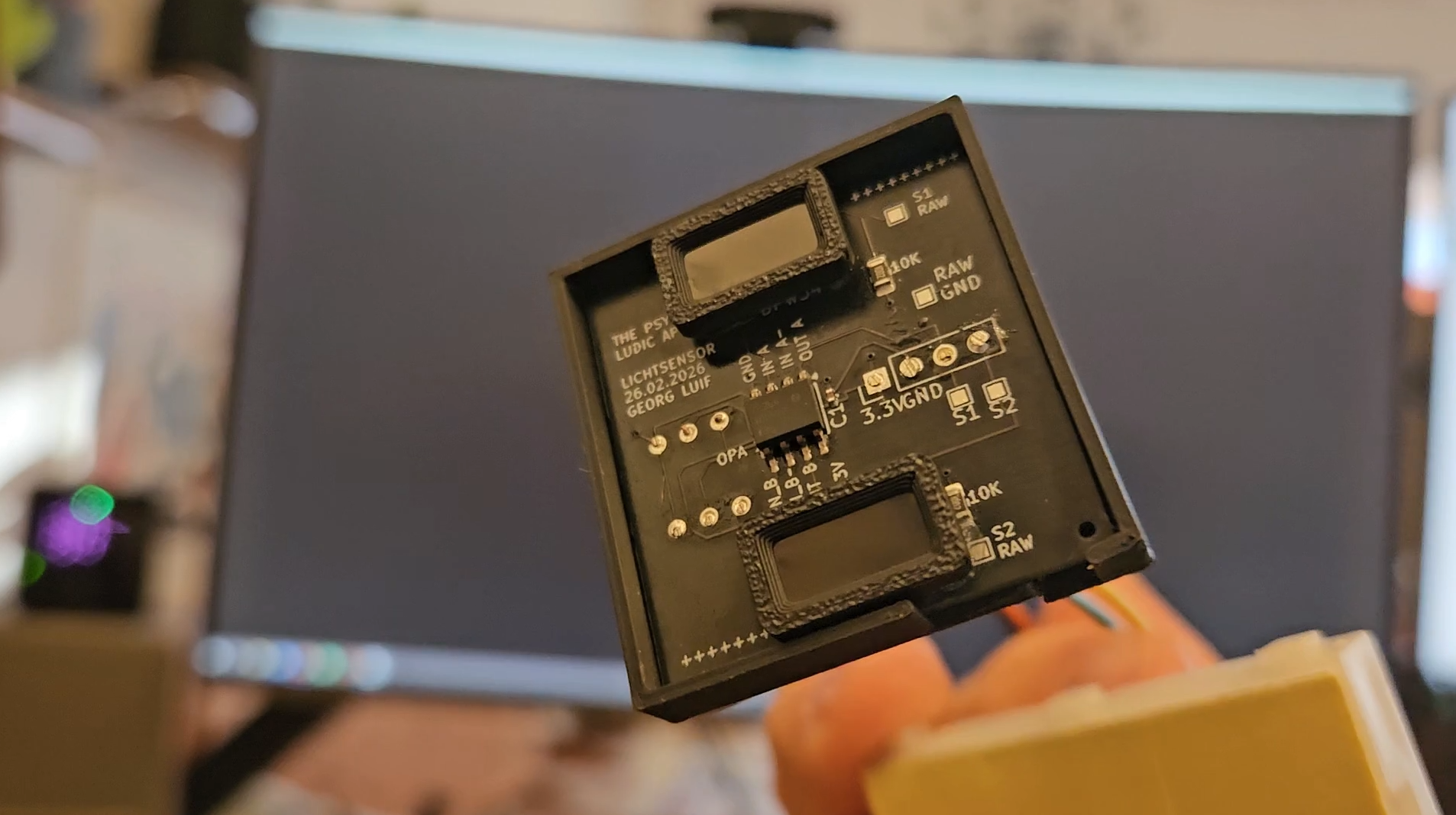 A hand holds a small black custom PCB with two chips in front of a computer monitor.
