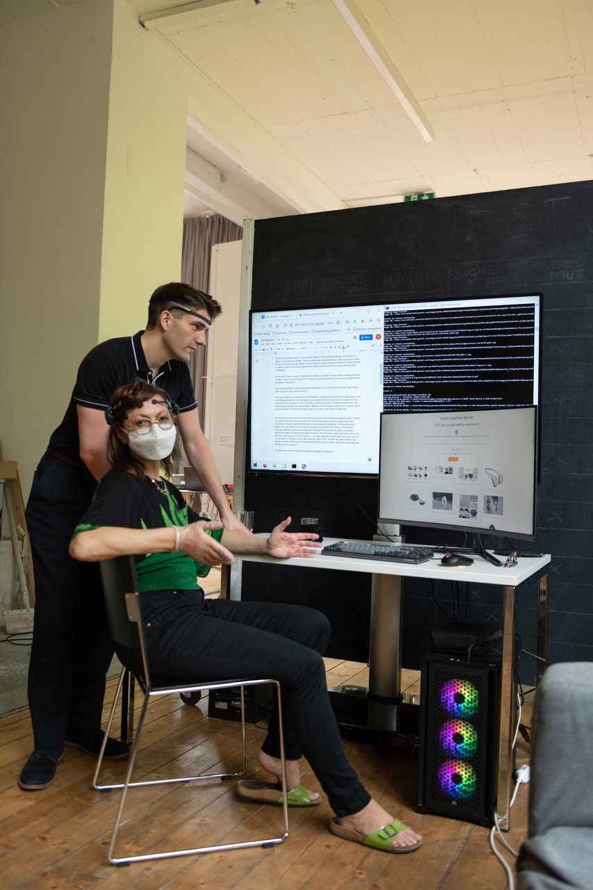 Two people stand by a large monitor and glowing desktop tower beside a seated participant wearing a mask and headband.