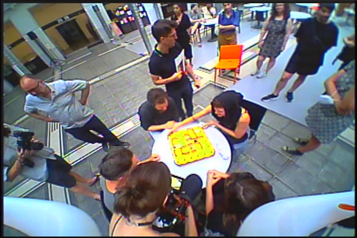 People around a round table play a board game in a bright indoor space with chairs and surrounding onlookers.