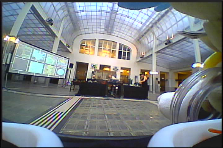 Low-angle view from a small drone over a tiled hall toward a central table and wall of screens under a glass roof.