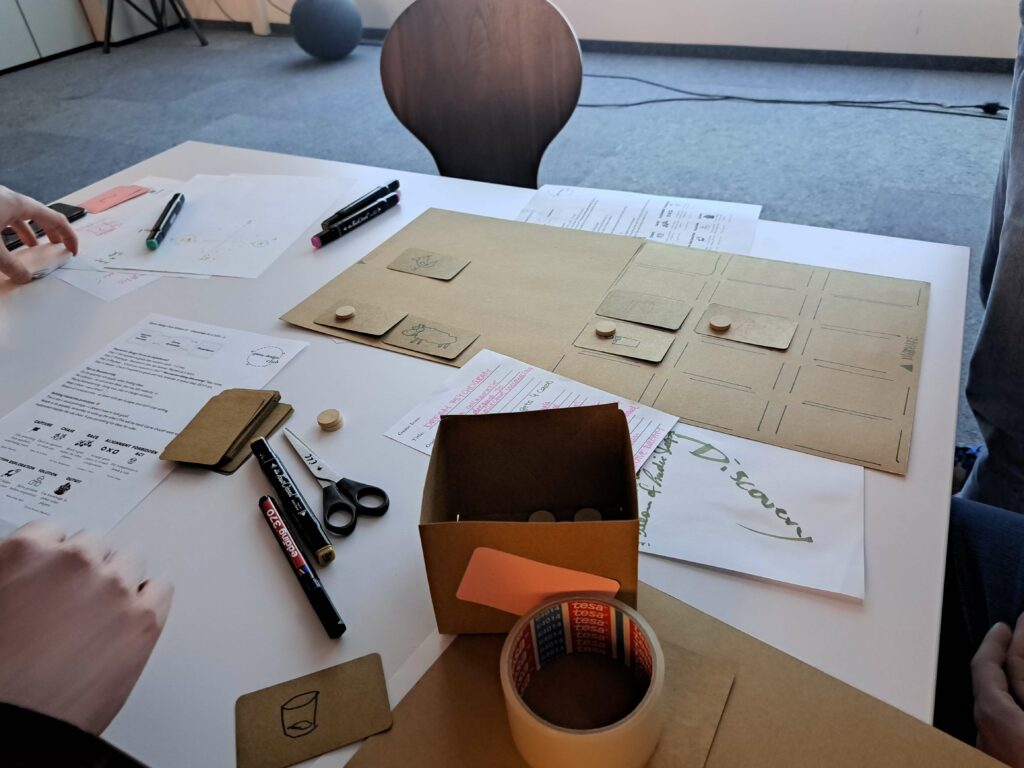 Hands and tools surround paper game prototypes, scissors, markers, and an open box on a tabletop.