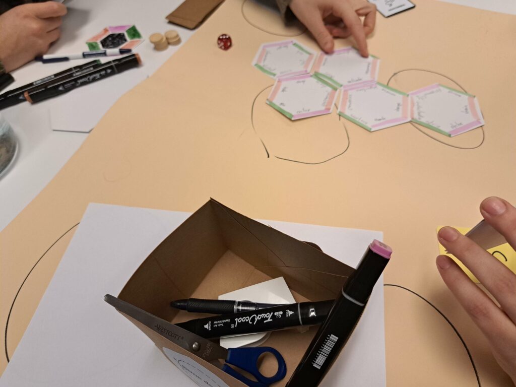 Hands draw colored hexagon game pieces on paper around a table with markers and scissors.