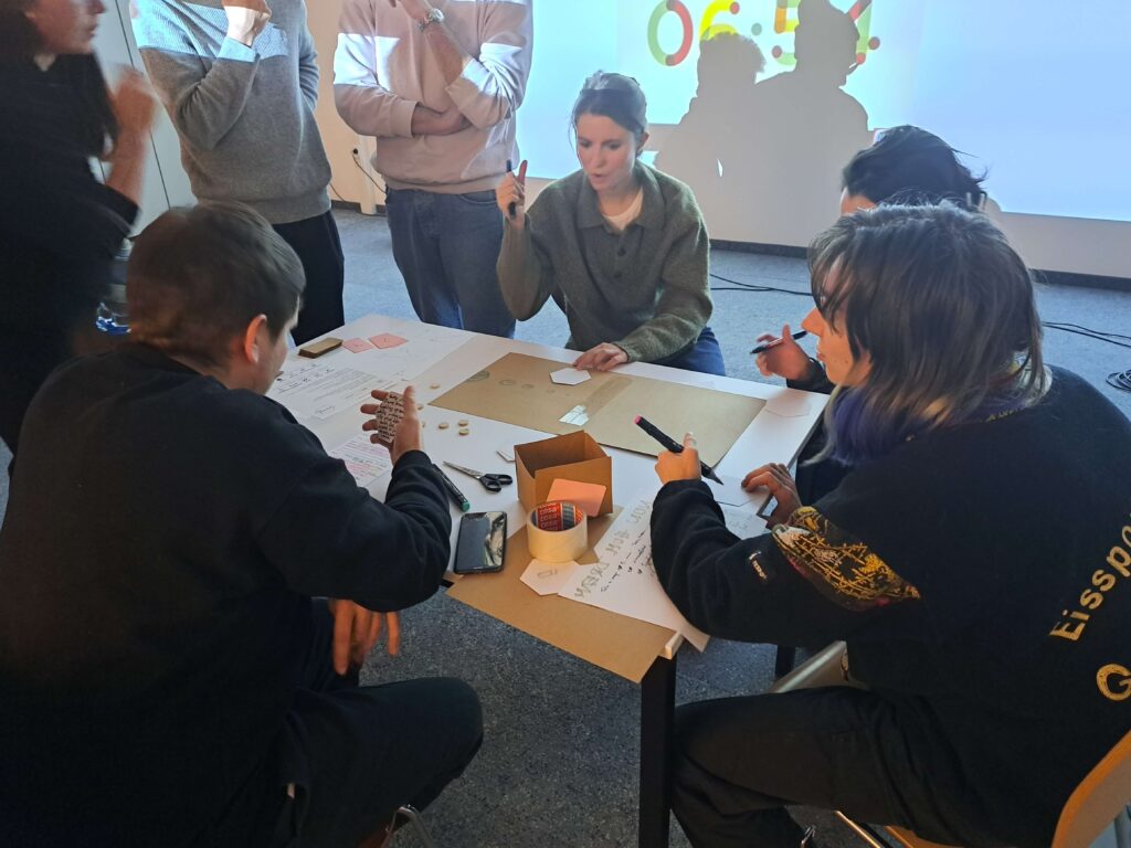 Students playtest a tabletop game prototype around a table in a classroom.