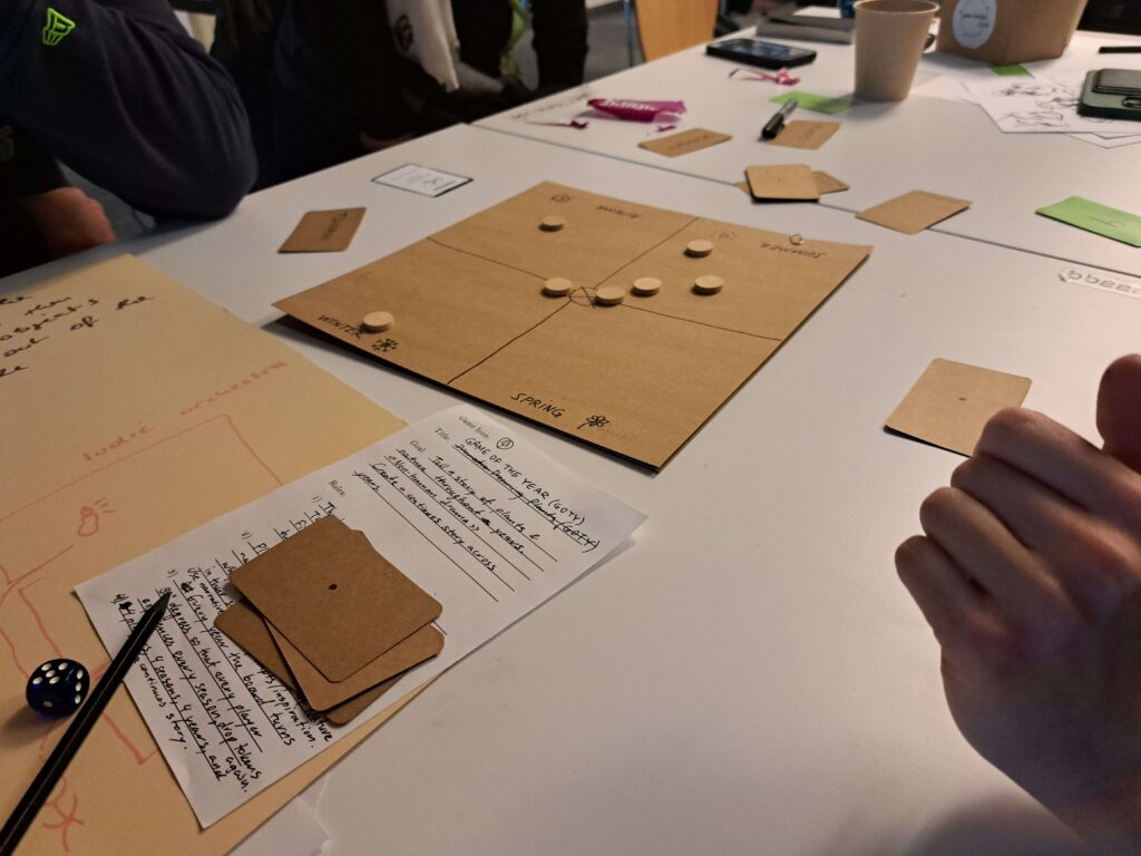 Hands and game cards around a cardboard board with tokens and handwritten notes on a table.