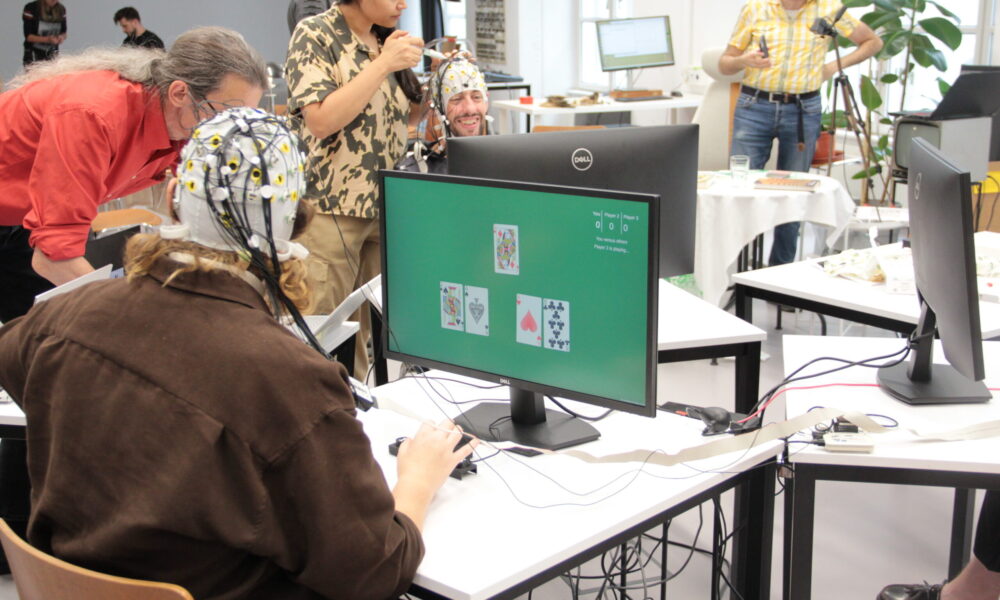 People with EEG caps gather around a monitor showing card-like game screens in a workshop setting.