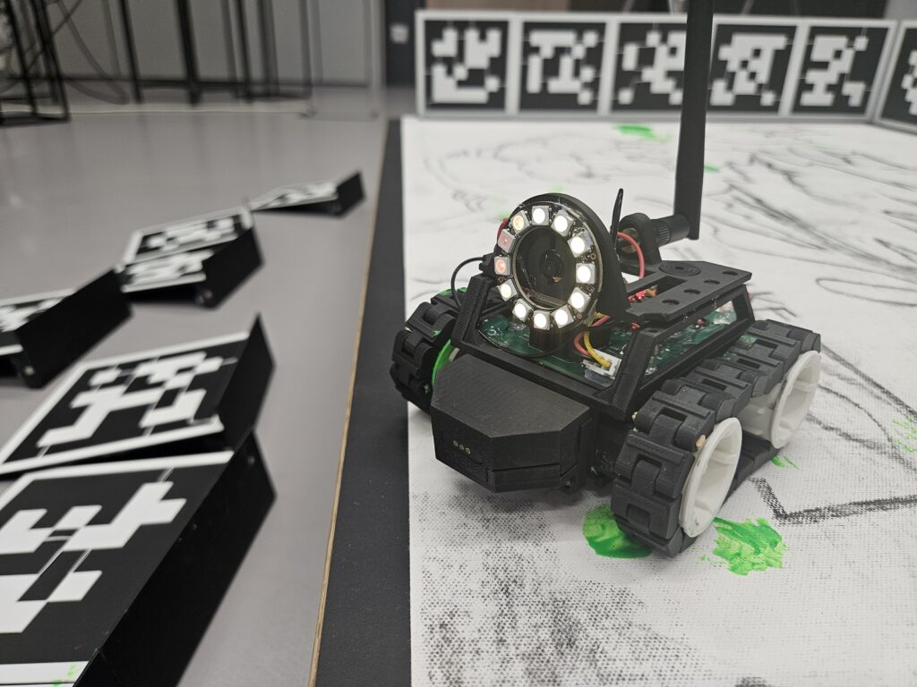 Small tracked robot with a circular LED ring sits on paper beside scattered black-and-white square markers.