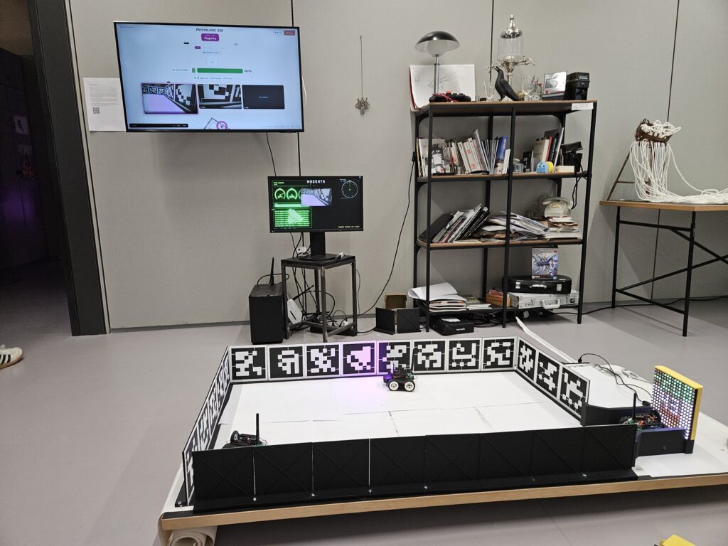 Three small robots and a charging station sit inside a white-bordered arena in a workshop.