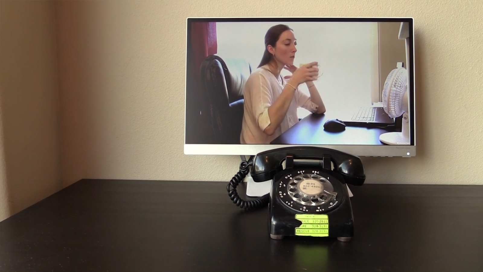 Black rotary phone on a desk in front of a monitor showing a woman speaking.