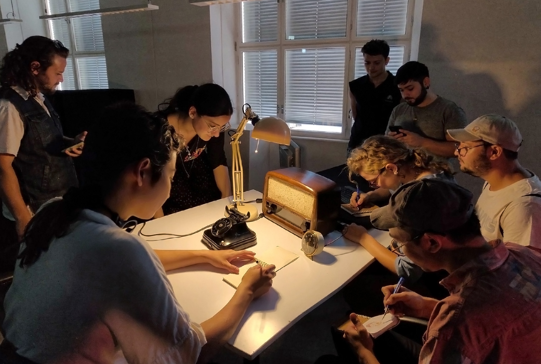 People gathered around a table with a vintage rotary telephone and lamp, taking notes in a dim room.